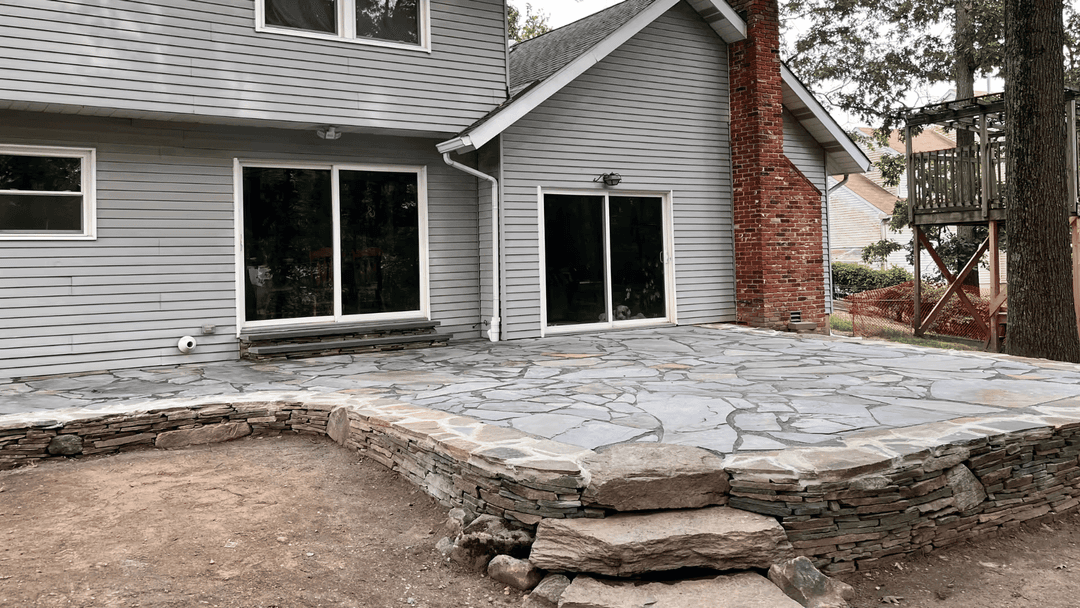 Residential patio made of stone, featuring a gray house and large glass doors.