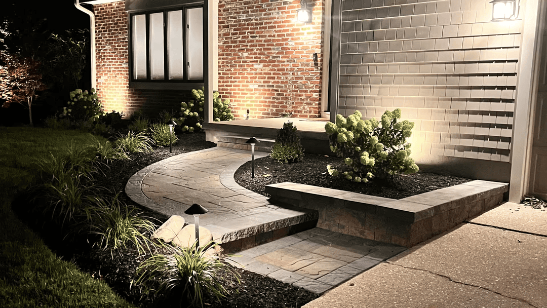 Well-lit driveway with paver stones, lush greenery, and modern landscape lighting at night.