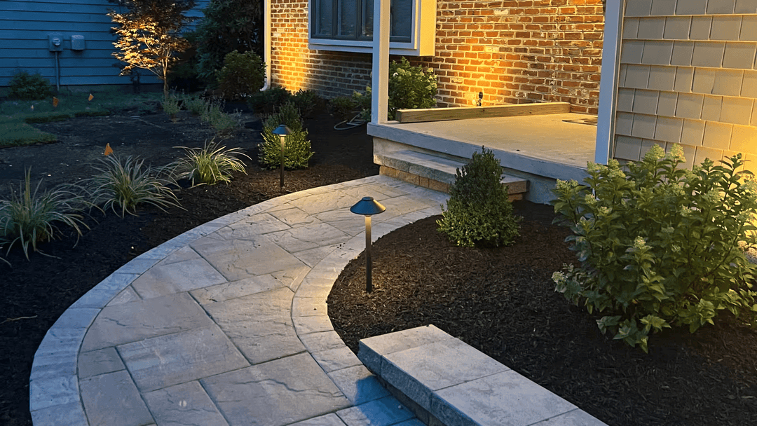 Illuminated walkway with landscaping near a brick house entrance at night.