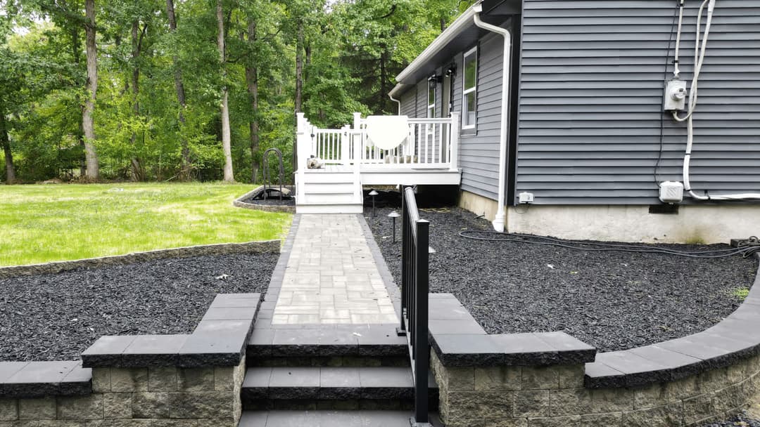 Backyard patio with gray stone walkway, black mulch, and wooden deck surrounded by trees.