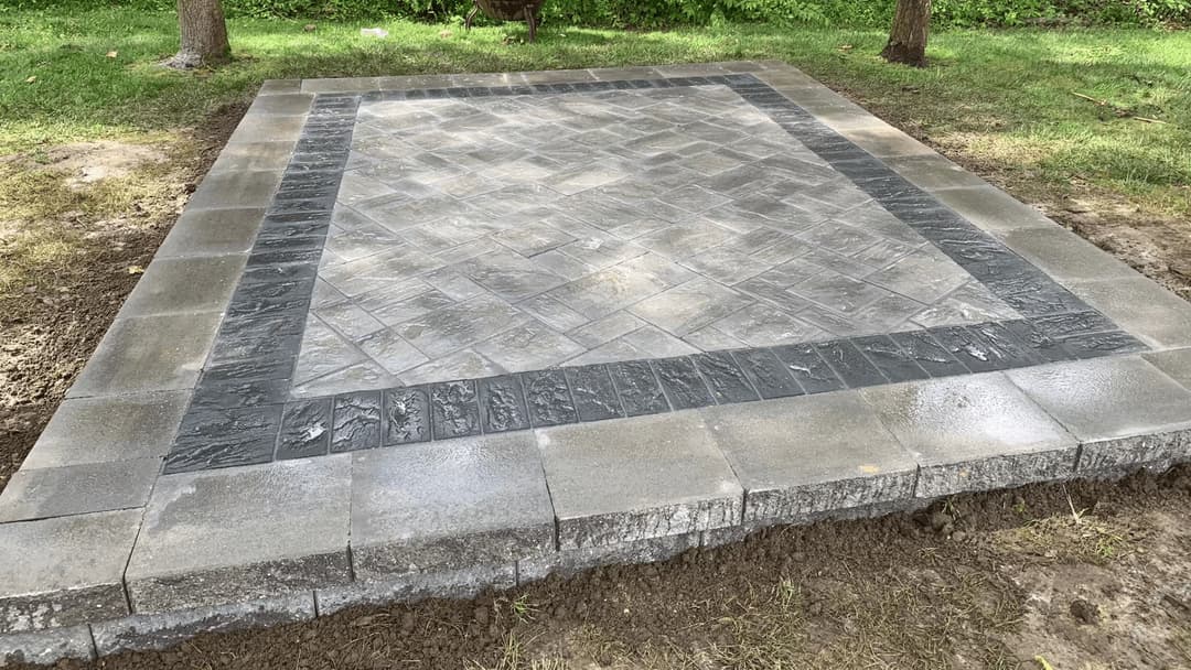 Paver patio installation with interlocking stones and patterned border in a grassy area.