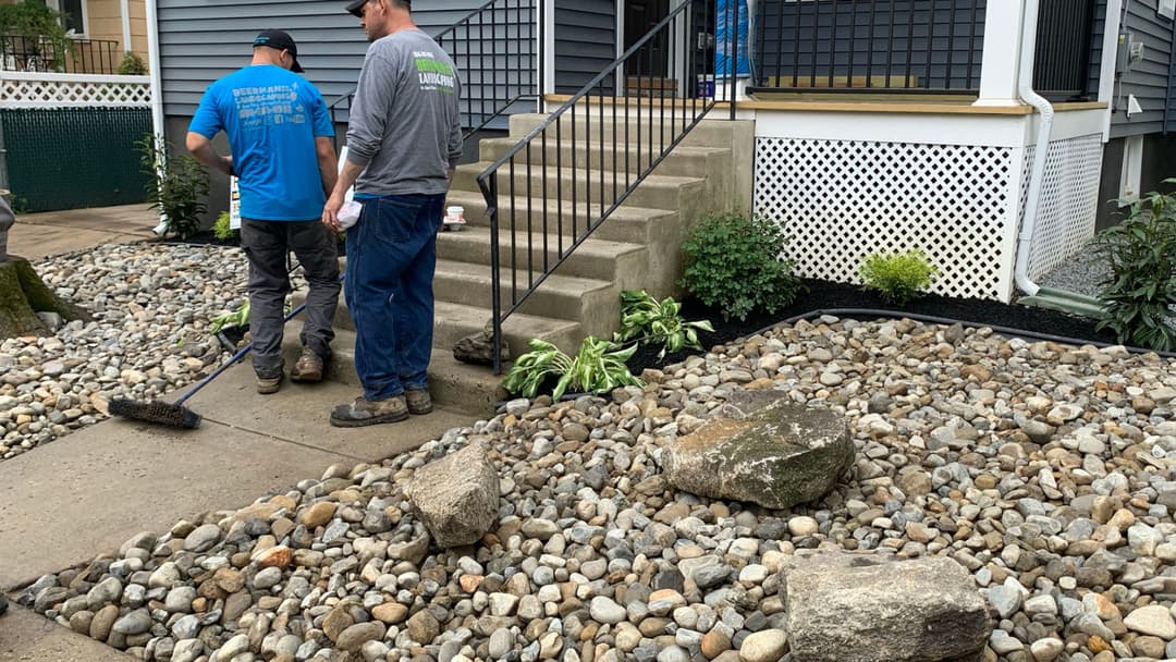 Two landscape workers arranging stones and plants near a home's entrance.