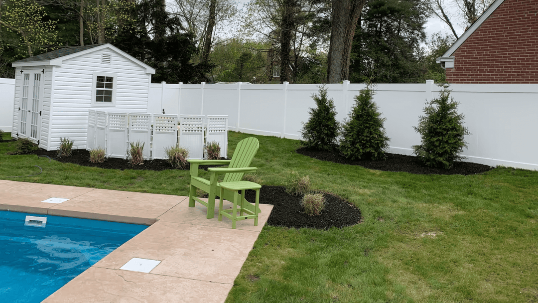 Backyard with a pool, green chair, white shed, and landscaped planting beds near a white fence.