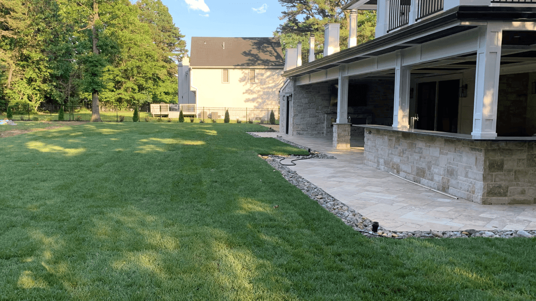 Spacious backyard with green lawn, stone patio, and nearby houses under blue sky.