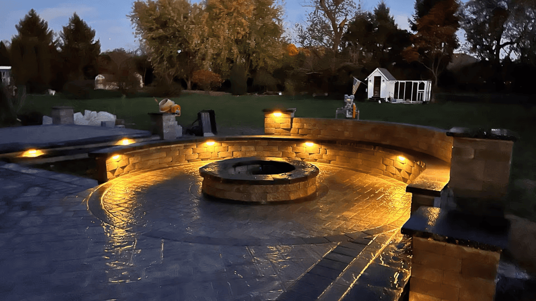 Outdoor fire pit surrounded by stone seating and illuminated landscape lighting at dusk.