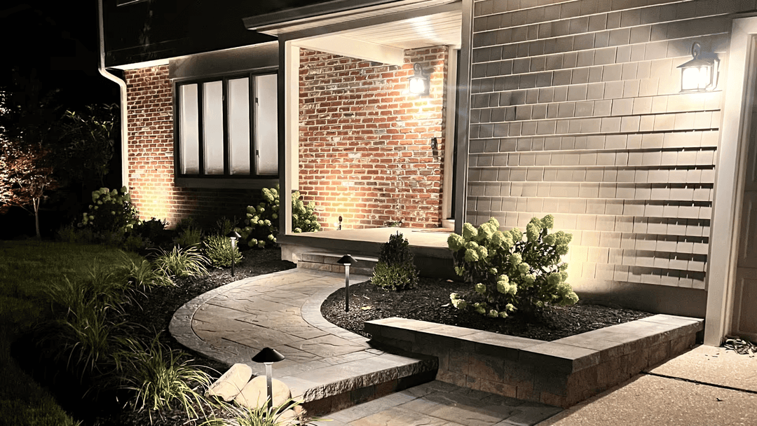 Modern home entryway with illuminated pathway, stone seating, and lush landscaping at night.