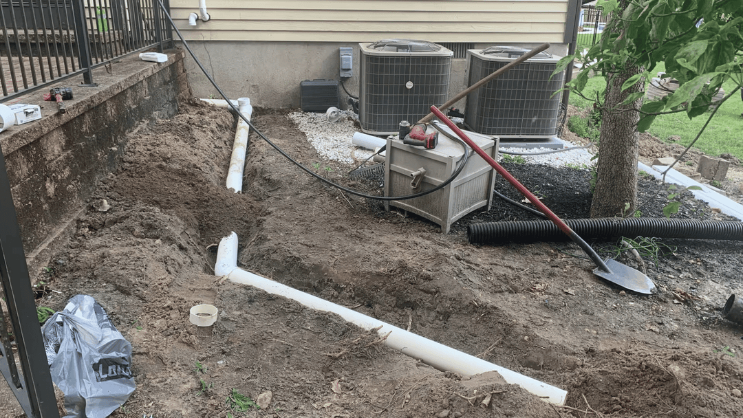 Excavated yard showing drainage pipes, tools, and air conditioning units in background.