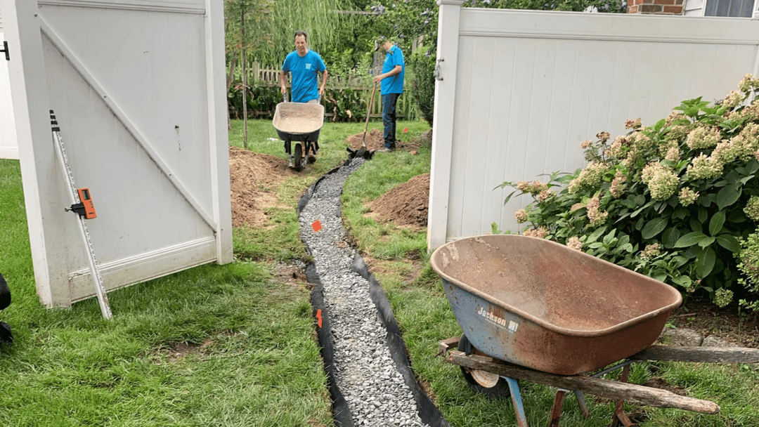 Workers laying gravel pathway in a residential yard with wheelbarrows and tools.
