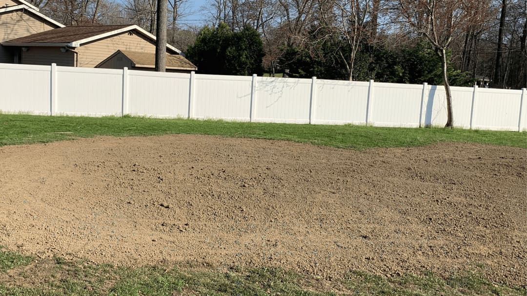 Bare soil area in backyard with white vinyl fence and trees in the background.