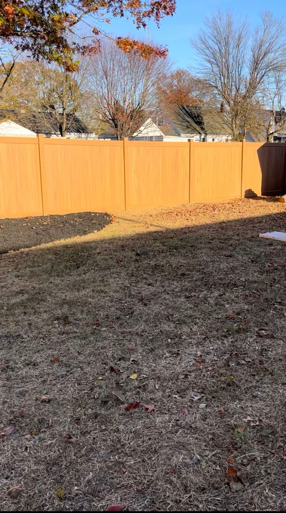 Wooden fence in sunny backyard with leaf-covered ground and bare trees in autumn.