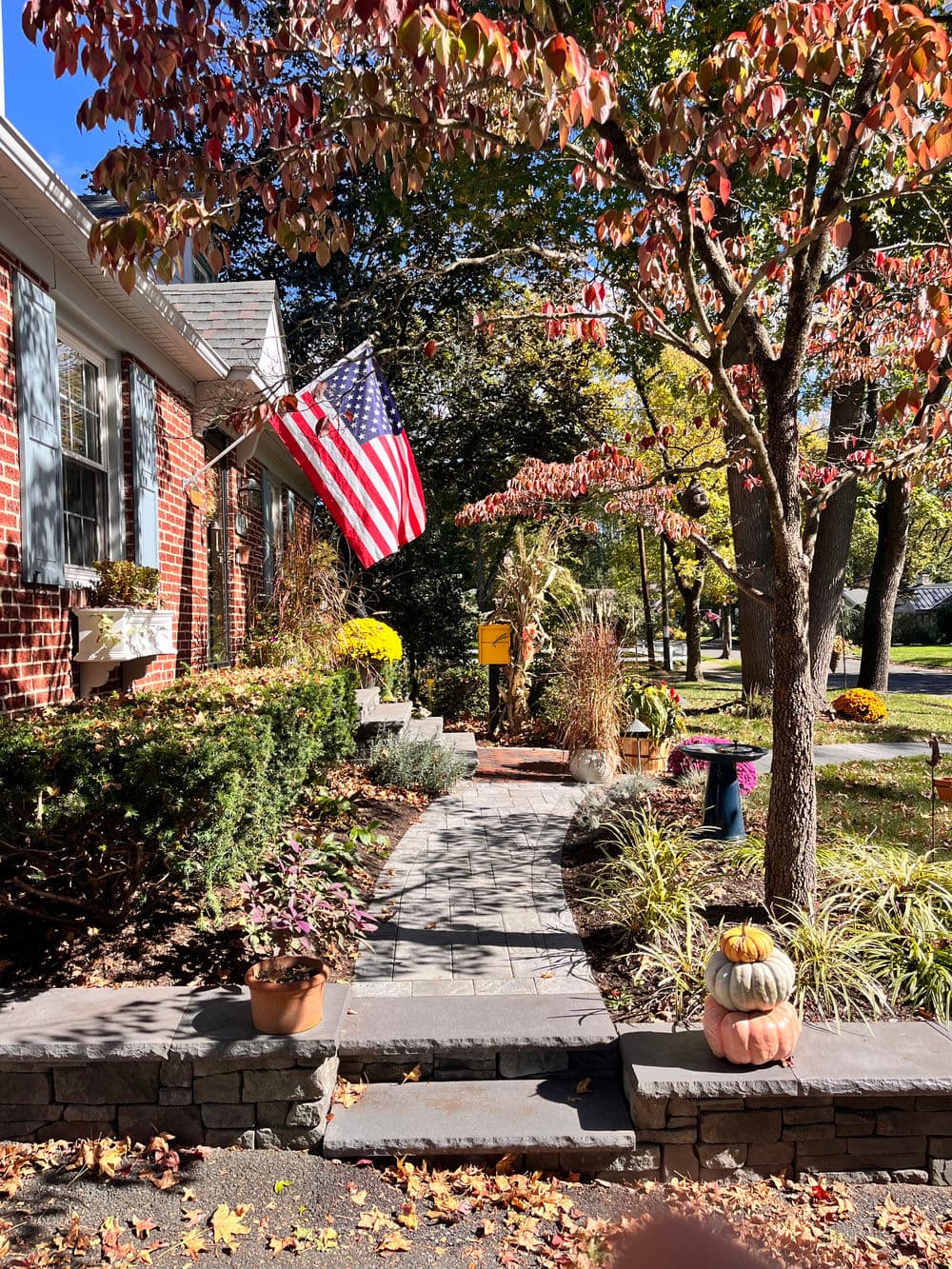 Autumn scene with American flag, pumpkins, and colorful foliage lining a stone walkway.
