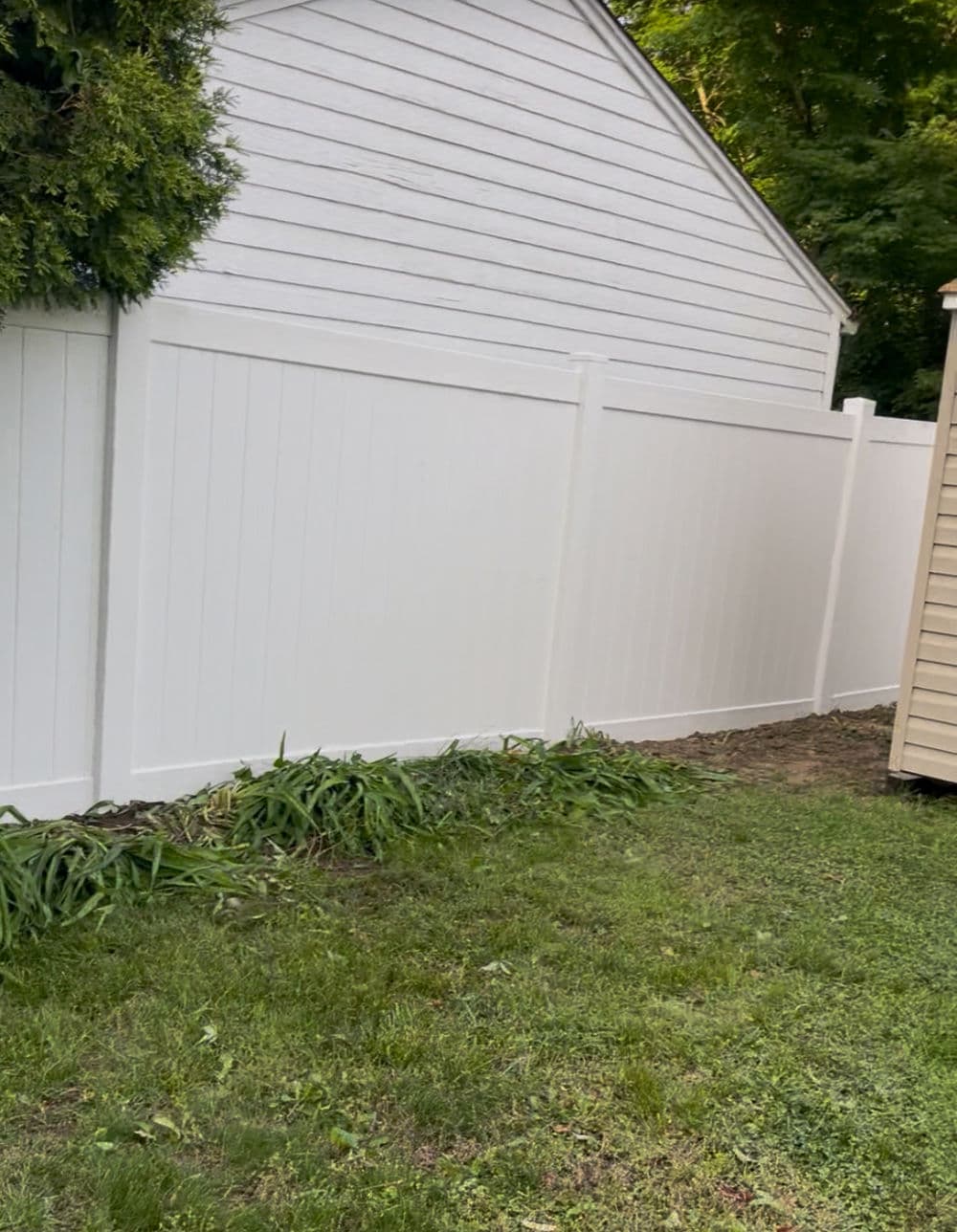 White vinyl fence alongside a grassy area and a shed against a backdrop of trees.