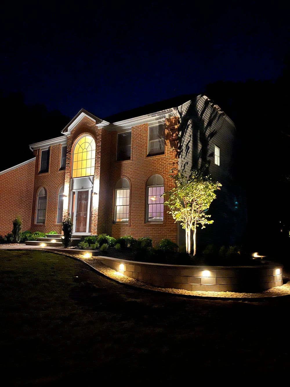 Illuminated brick house exterior at night with landscaped front yard and decorative lighting.