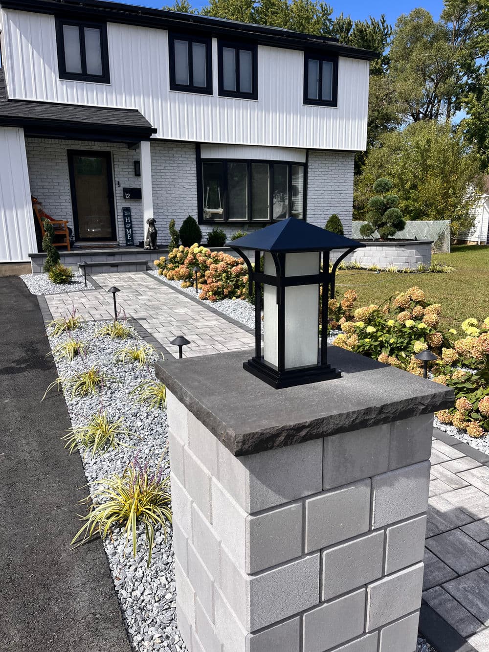 Modern outdoor lantern on a stone pillar in front of a contemporary home with landscaped garden.