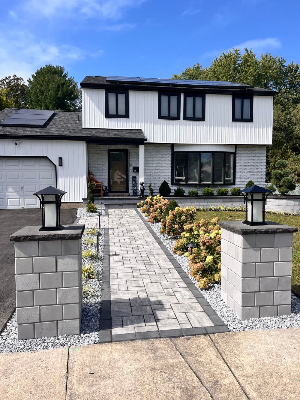 Modern white and gray house with solar panels, landscaped yard, and paved walkway.