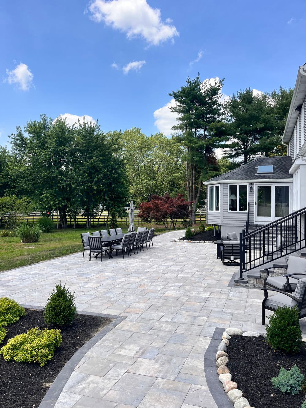 Spacious patio area with stone pavers, surrounded by green trees and plants on a sunny day.