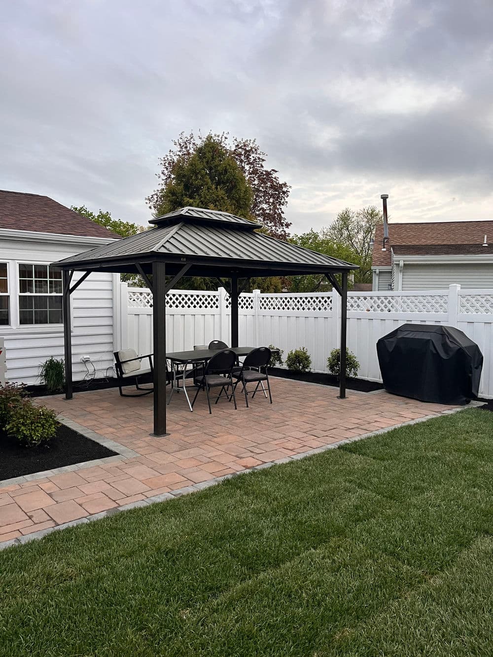 Backyard gazebo with seating area, brick patio, and grill under a cloudy sky.