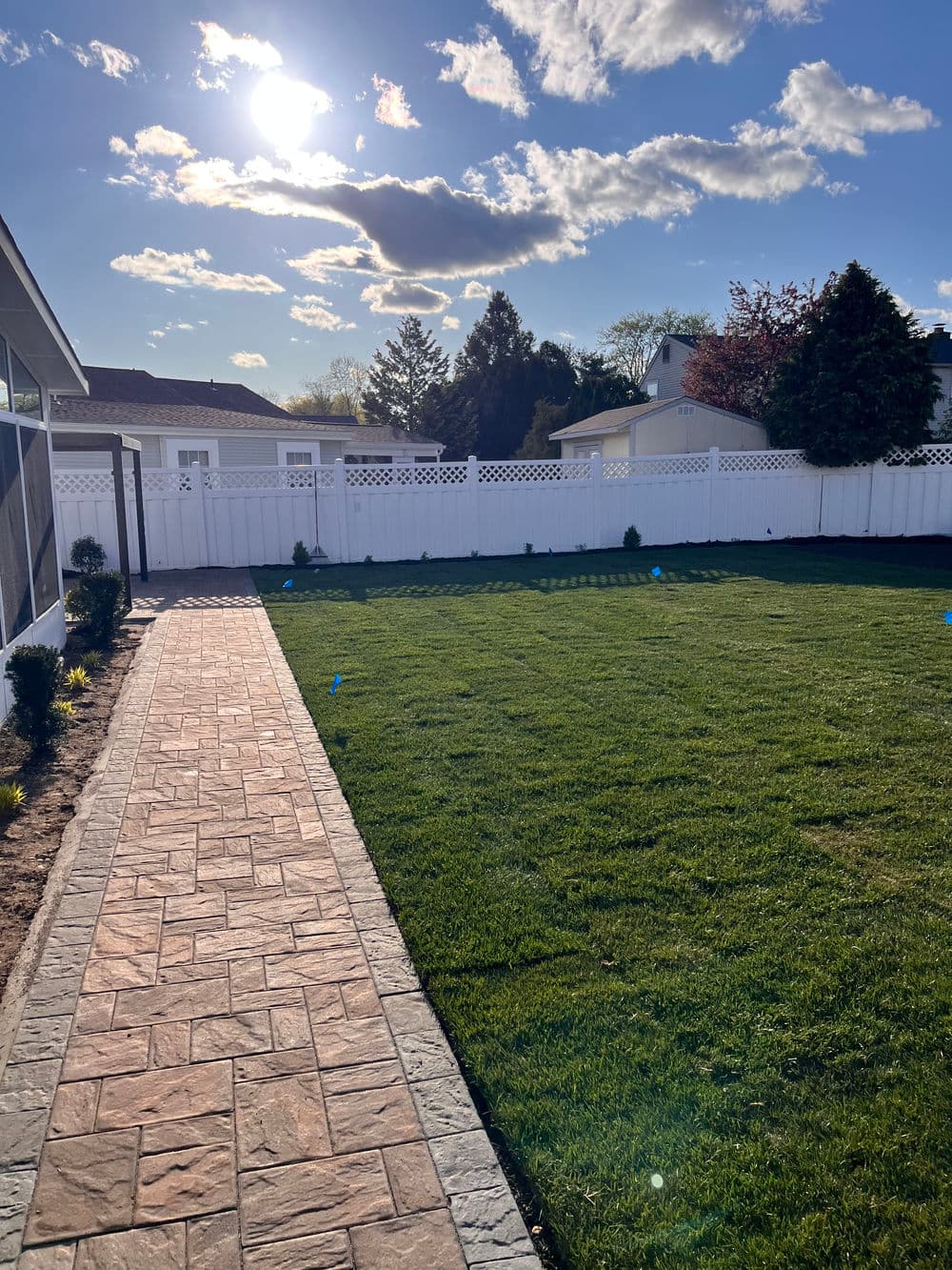 Sunny backyard with freshly mowed grass, paved pathway, and white picket fence.