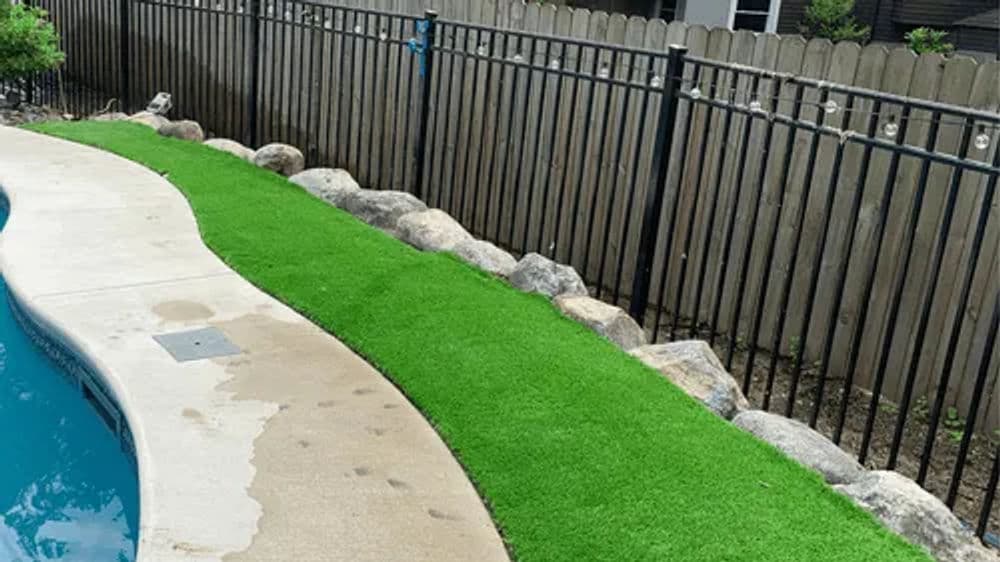 Artificial grass pathway beside a pool, bordered by rocks and a black fence.